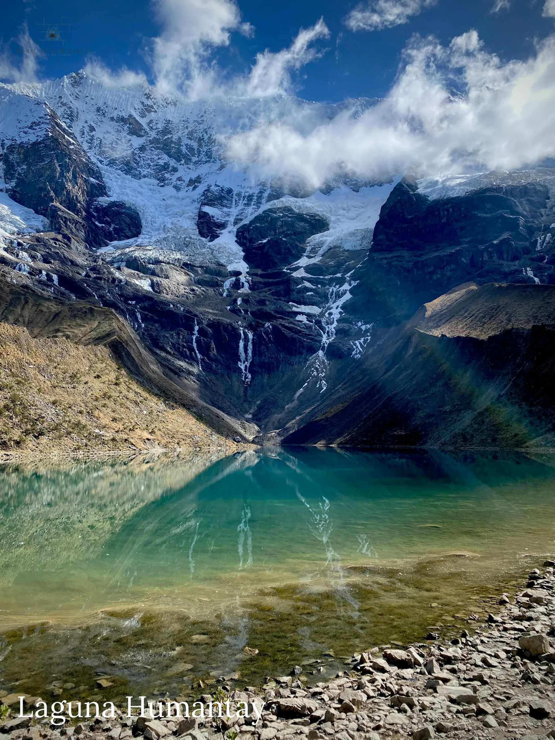 Águas cristalinas turquesa-esmeralda da Lagoa de Humantay vistas da margem rochosa com reflexo perfeito do nevado e geleiras descendo — a 4.180m de altitude na Cordilheira Vilcabamba, Cusco, Peru
