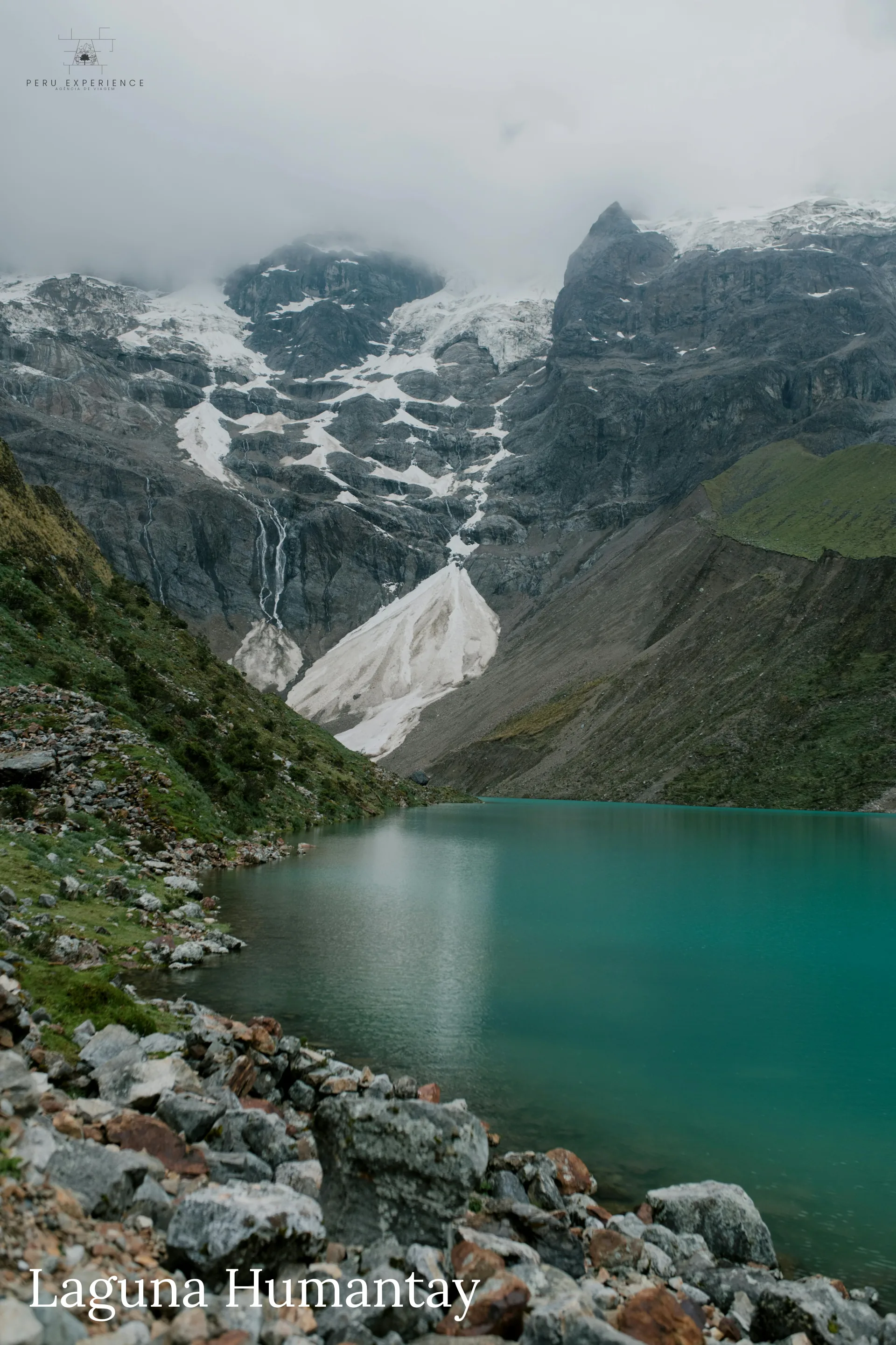 Margem rochosa da Lagoa de Humantay com vista do anfiteatro natural formado pelas montanhas e geleiras — perspectiva do trekker após 1h20 de subida desde Soraypampa, Cusco, Peru