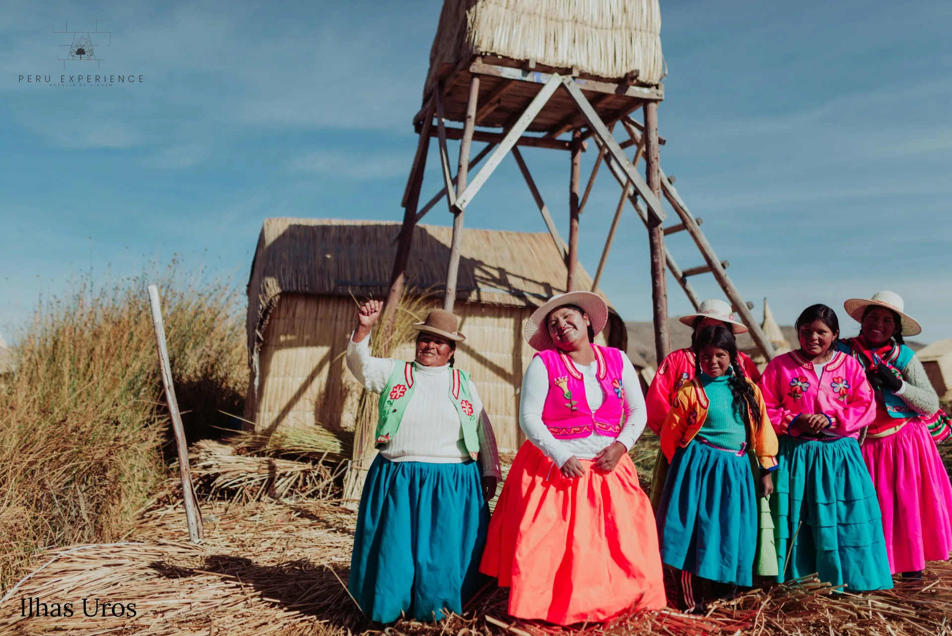 Mulheres Uros sorrindo em trajes tradicionais coloridos (saias azuis e laranjas, coletes bordados, chapéus típicos) em frente a torre de observação de totora no Lago Titicaca — comunidade pré-inca viva em Puno, Peru