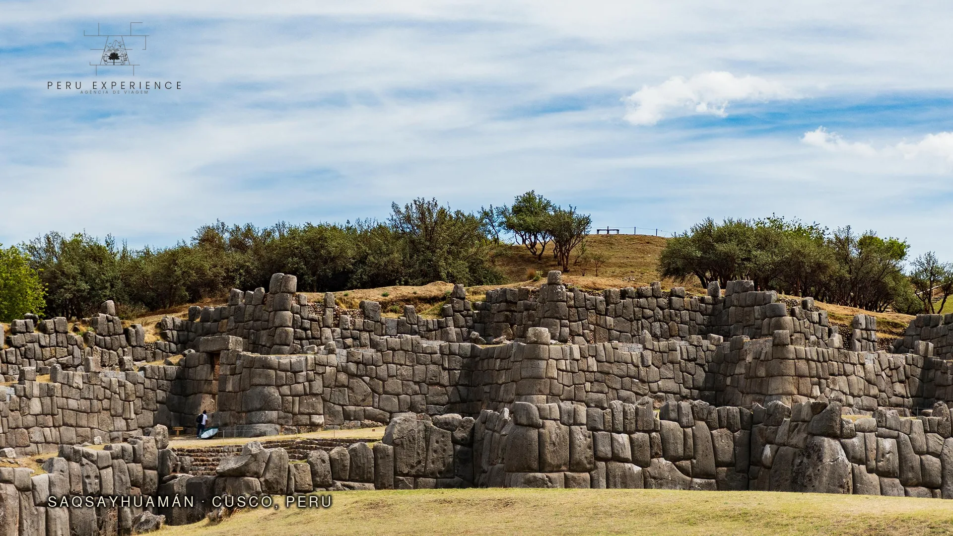 Sacsayhuaman em Cusco — muralha megalítica em zigue-zague da fortaleza inca formada por três plataformas sobrepostas representando os dentes do puma sagrado, com rochas de até 120 toneladas encaixadas com precisão milimétrica sem argamassa, vista frontal de uma pessoa próxima às pedras para escala monumental