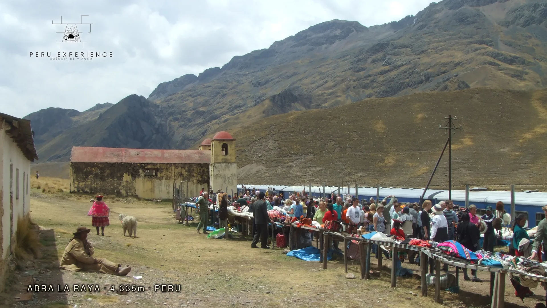 Paso de La Raya a 4.335 metros de altitude — ponto mais alto da rota Cusco-Puno com mercado de artesãos quechuas vendendo têxteis de alpaca, trem turístico Andean Explorer estacionado e alpaca caminhando entre as barracas, limite geográfico entre Cusco e Puno