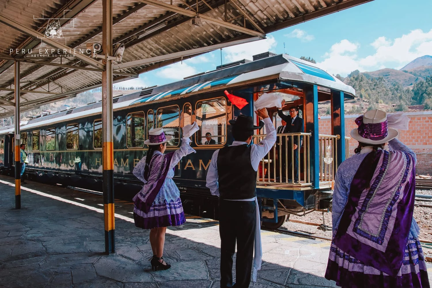 Dançarinos com trajes tradicionais recebem passageiros ao lado do trem Hiram Bingham.